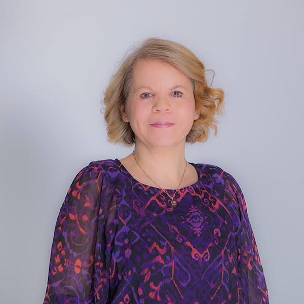 Portrait of a smiling woman with short blond hair in a purple patterned blouse against a light gray background.