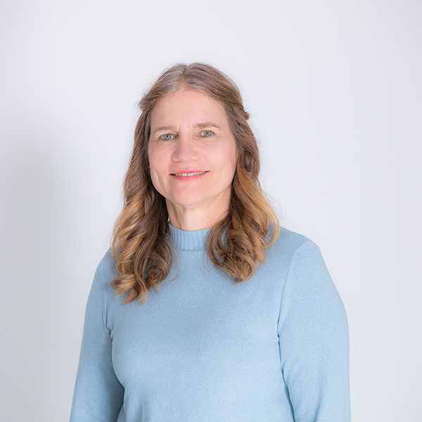 Smiling woman with wavy light brown hair in a blue sweater, posing against a pale background (head-and-shoulders portrait).
