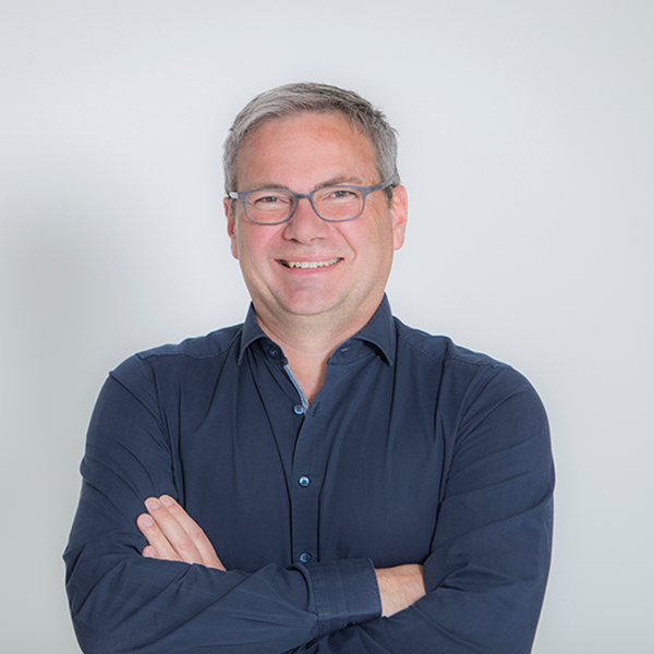 Smiling middle-aged man with gray hair and glasses, wearing a navy button-down shirt, arms crossed against a light background.