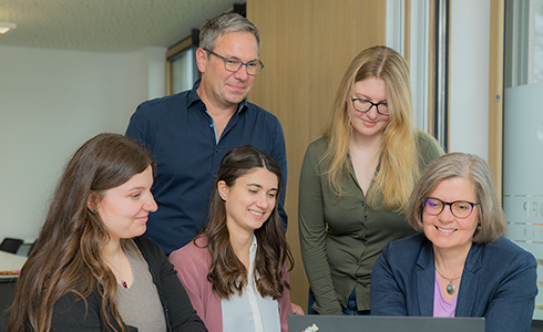 Five colleagues gathered around a laptop, collaborating in a bright office space by a window