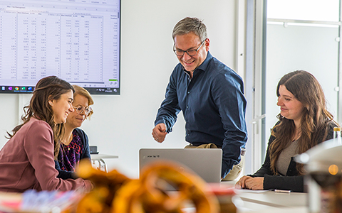 A man in a blue shirt explains something to three colleagues around a laptop in a bright office while a spreadsheet is visible on a wall screen.