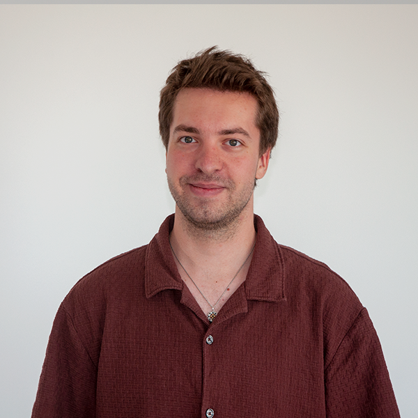 Smiling man with short brown hair wearing a maroon button-down shirt against a pale gray wall.