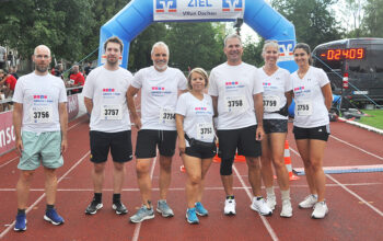 Group of seven runners in white shirts posing at the finish line under a blue inflatable arch on a red track path.