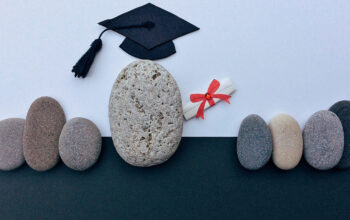 Graduation cap rests on a large pebble with a small rolled diploma tied in red ribbon among smooth stones.