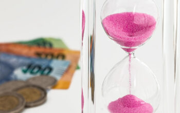 Pink sand hourglass in clear glass with coins and currency in the blurred background, conveying time and money symbolism.