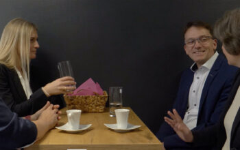 Group of four adults seated at a wooden table, chatting over coffee, with a basket of pink napkins nearby.