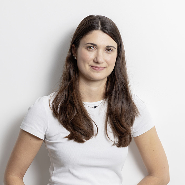 Young woman with long brown hair wearing a white t-shirt, standing against a white wall and smiling subtly.
