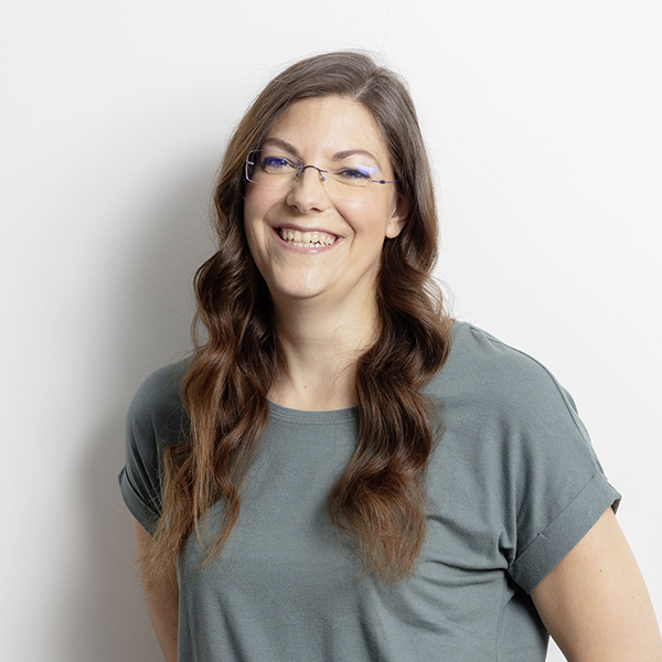 Smiling woman with long wavy brown hair and blue glasses wearing a gray t-shirt against a white wall.