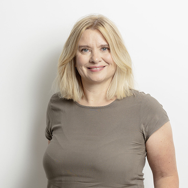 Smiling blonde woman wearing a taupe shirt, looking at the camera against a white background.