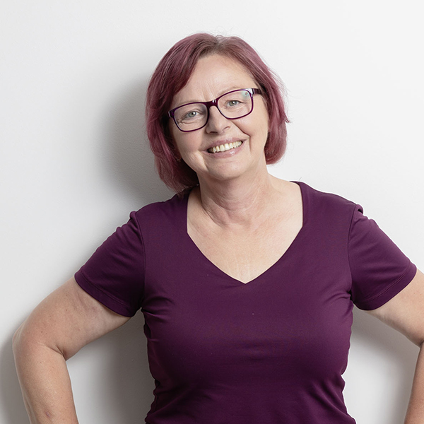 Smiling woman with short purple hair and glasses posing with hands on hips against a white wall in a purple V-neck shirt.
