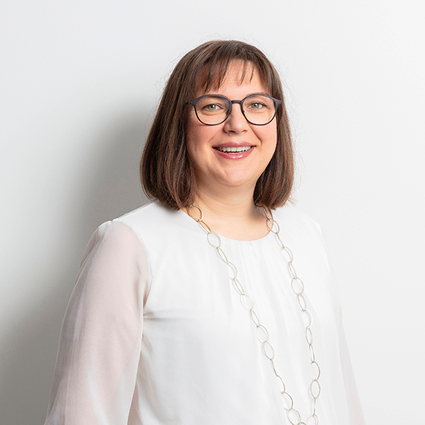Portrait of a smiling woman with short brown hair and glasses, wearing a white blouse and long circular necklace, standing against a light wall