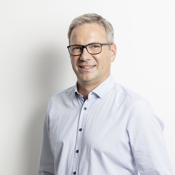 Smiling middle-aged man with gray hair and glasses in a light blue button-down shirt against a white background.