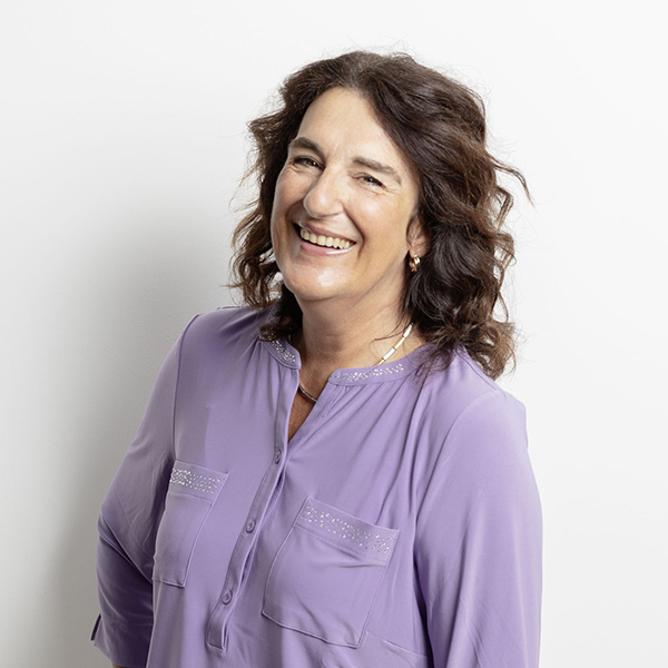 Smiling woman with wavy brown hair in a lavender blouse posing against a white background.