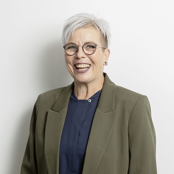 Smiling woman with short white hair and round glasses, wearing an olive blazer against a white wall.