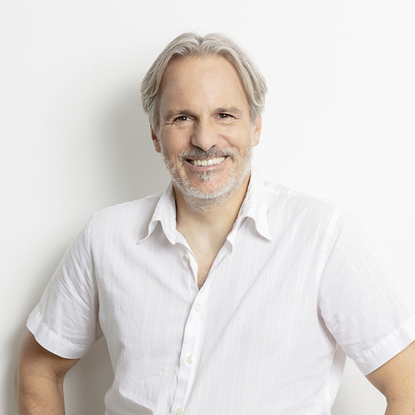 Smiling middle-aged man with gray hair and beard in a white short-sleeve button-down shirt, standing against a white wall.