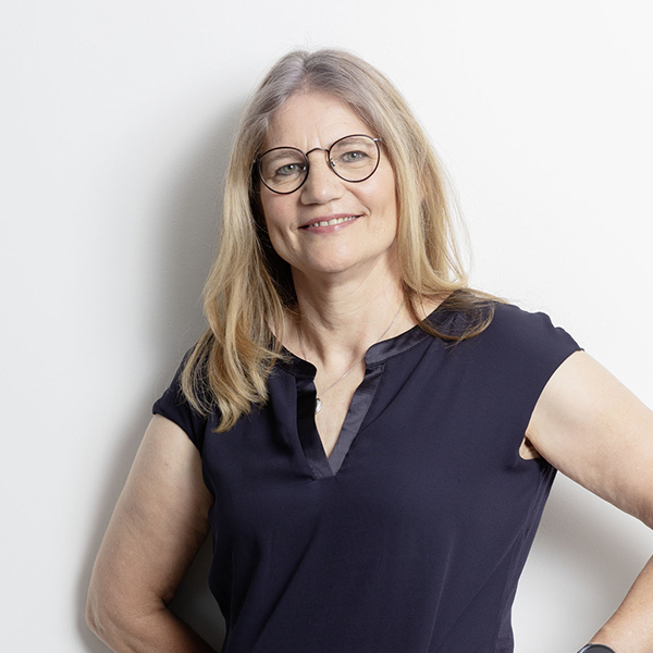 Smiling woman with round glasses and shoulder-length blond hair, wearing a navy blouse, posed against a white wall.