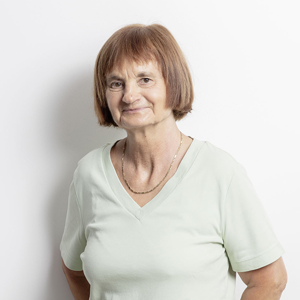 Smiling older woman with short light brown hair and a pale green V-neck shirt, standing against a white background.