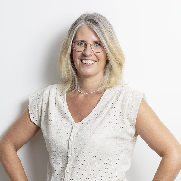 Smiling woman with gray-blonde hair and glasses, wearing a white eyelet blouse, against a white background.