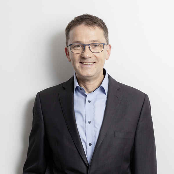 Smiling man in a dark suit and light blue shirt, wearing glasses, standing against a white wall (professional headshot).