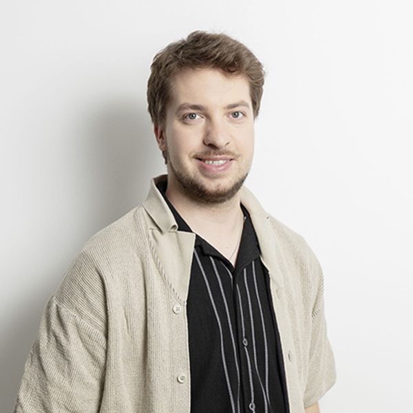 Portrait of a man with light brown hair and a beard, smiling at the camera, wearing a black striped shirt and beige cardigan against a white background.