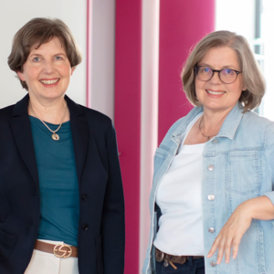 Two smiling women stand indoors beside pink columns; one wears a teal top with a navy blazer, the other a white top with a light denim jacket.