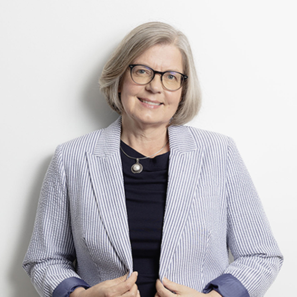 Portrait of an older woman with short gray hair and glasses, wearing a light striped blazer and dark top, smiling at the camera.