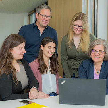 Five colleagues in an office look at a laptop together around a table, suggesting a collaborative meeting.