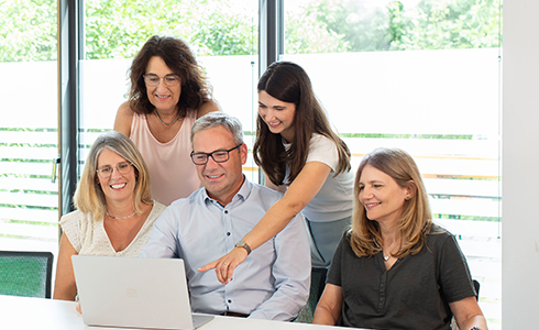 Five coworkers gathered around a laptop, with one woman pointing at the screen as others smile.